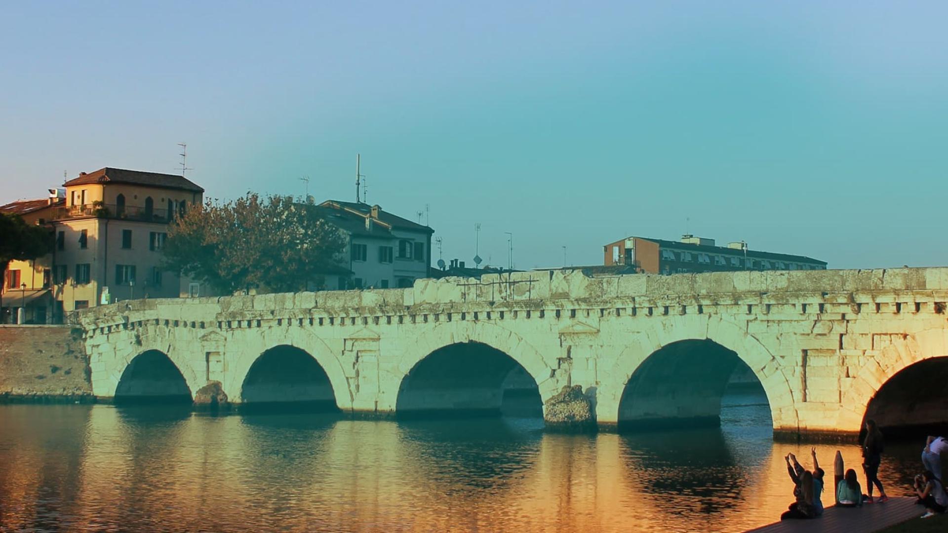 Ponte storico in pietra riflesso nell'acqua al tramonto, con persone sedute vicino.