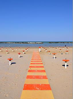 Spiaggia con passerella a righe e supporti per ombrelloni, cielo sereno.