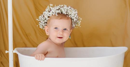 Bambino in vasca con corona di fiori bianchi, sfondo marrone chiaro.